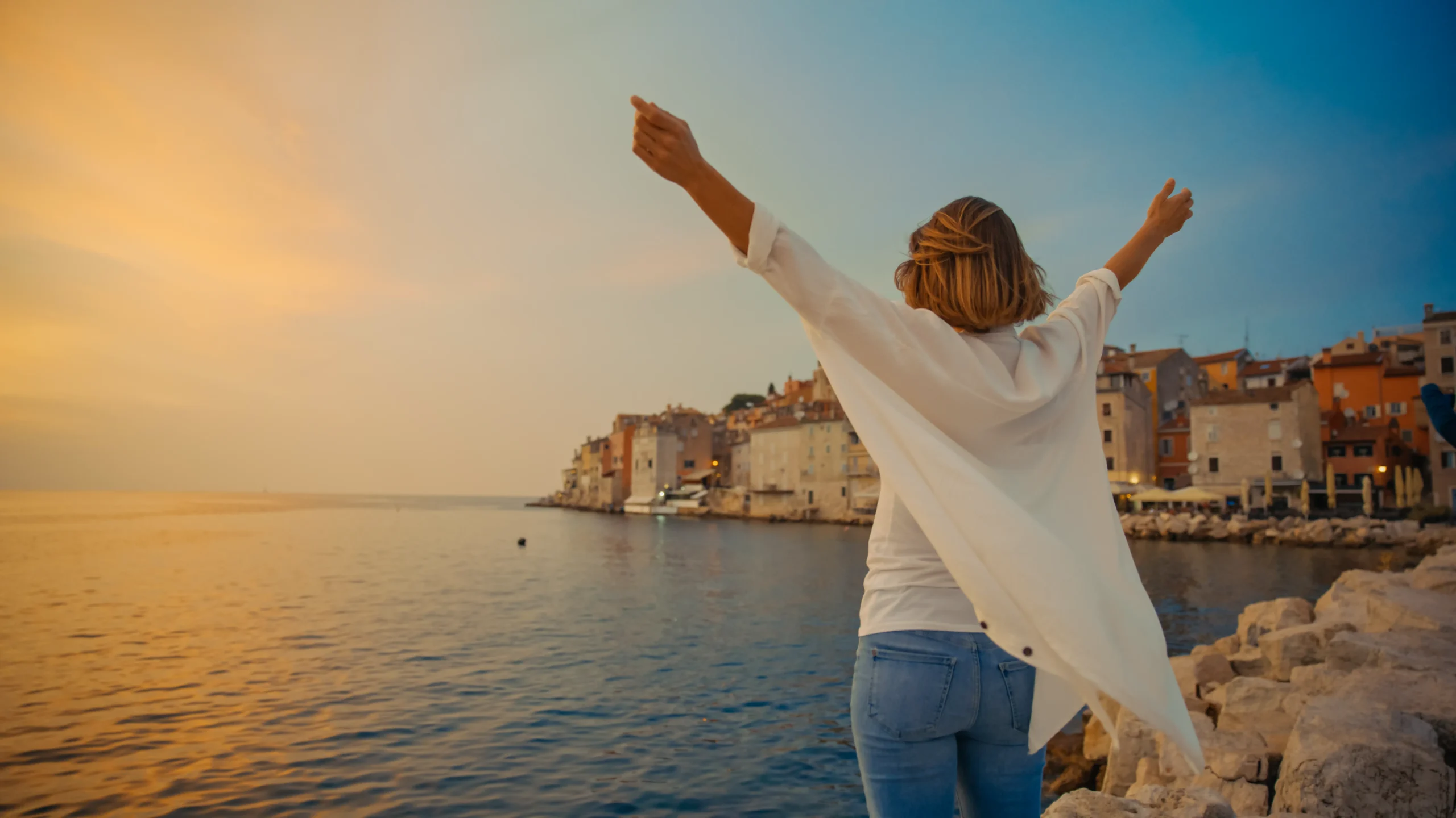 Woman standing on rocky shoreline with arms raised toward the sky, facing a golden sunset over the old town of Rovinj, Croatia