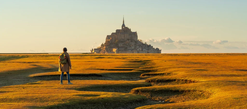 A traveller alone on the vast tidal flats of Normandy, standing still before Mont Saint-Michel — a moment of intention before the journey begins.