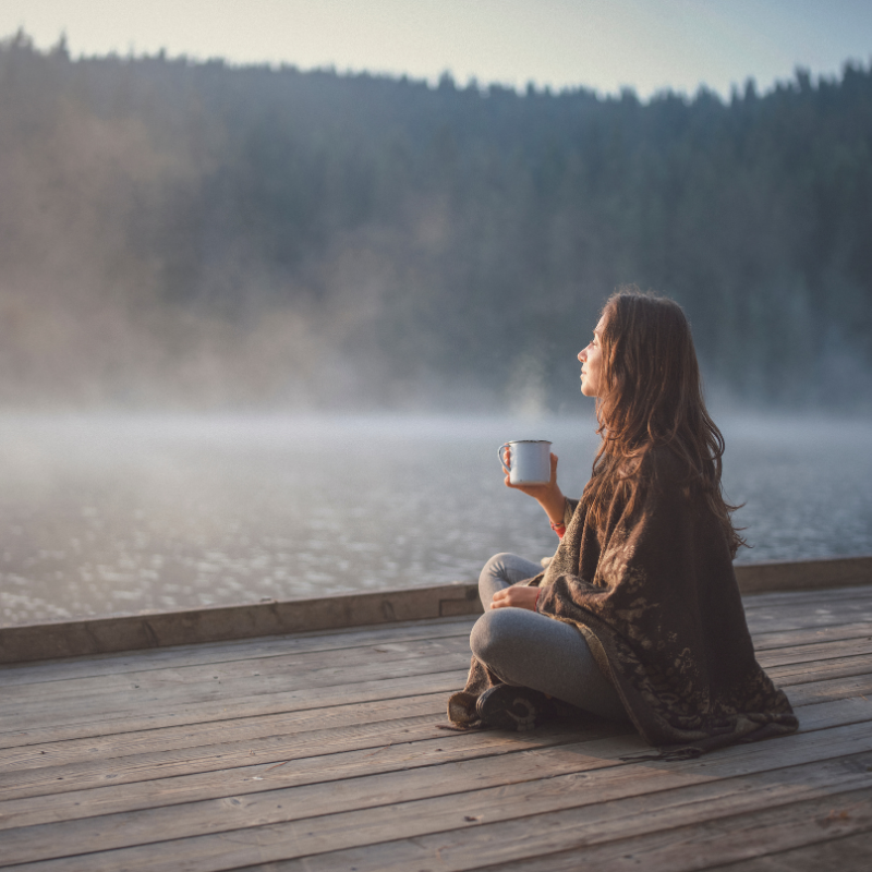 Woman wrapped in a blanket sitting cross-legged on a wooden dock, holding a steaming mug and looking out over a misty lake surrounded by forest at dawn.